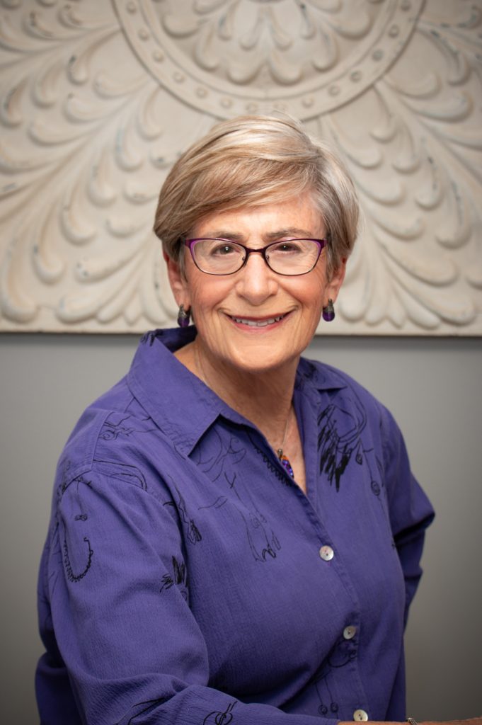 Senior woman smiling, wearing glasses, purple shirt, ornate background.